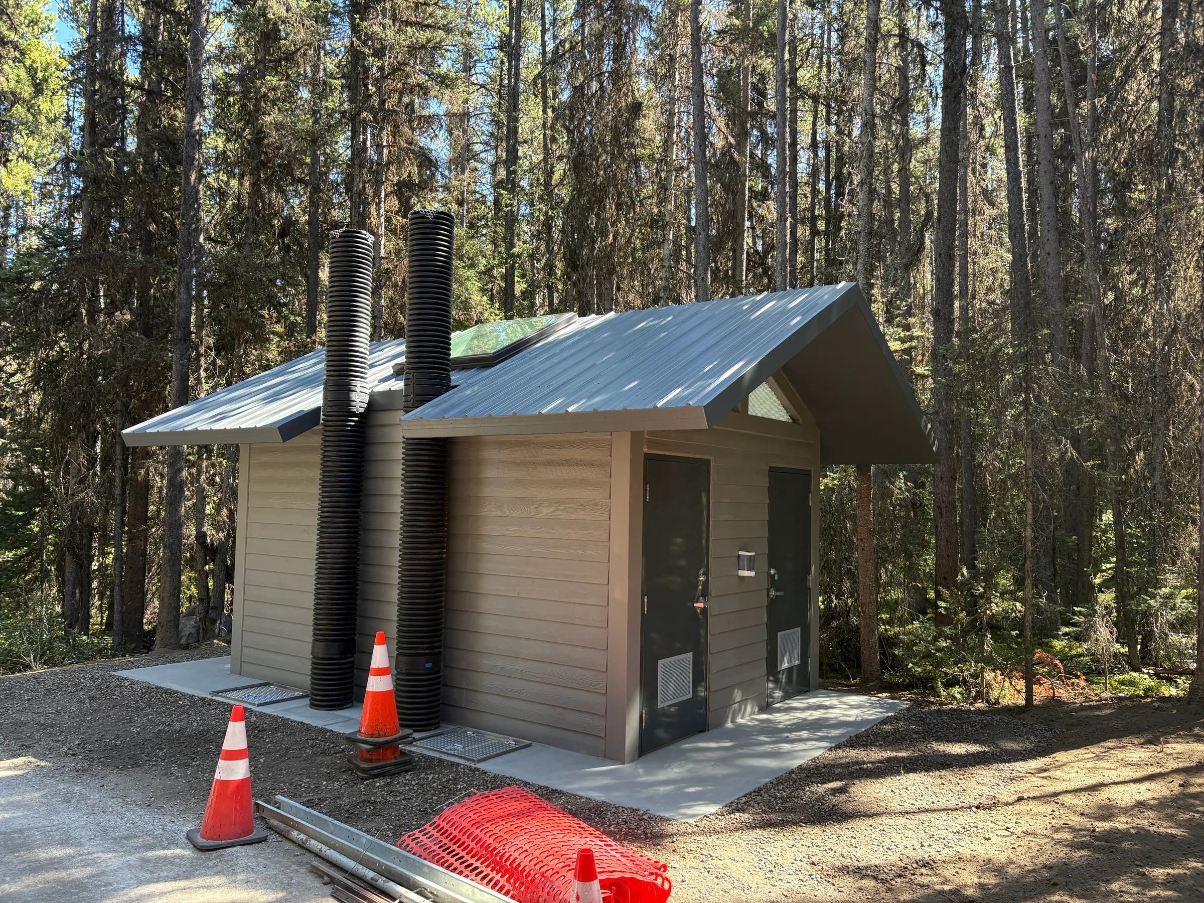 Side view of a new double vault toilet with metal roof and vent stacks in a pine forest at Kananaskis Region Vault Toilets, built by Premiere Builders