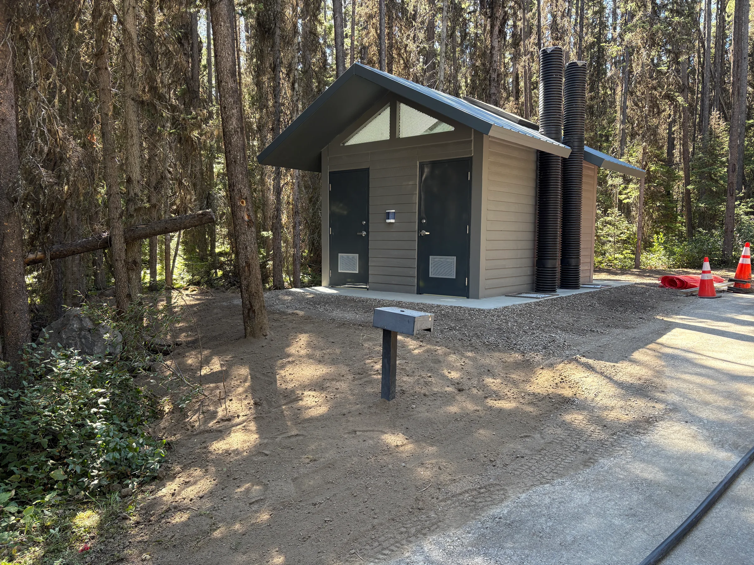 Completed vault toilet with grey siding, gabled roof and concrete apron set among tall pines at Kananaskis Region Vault Toilets, built by Premiere Builders