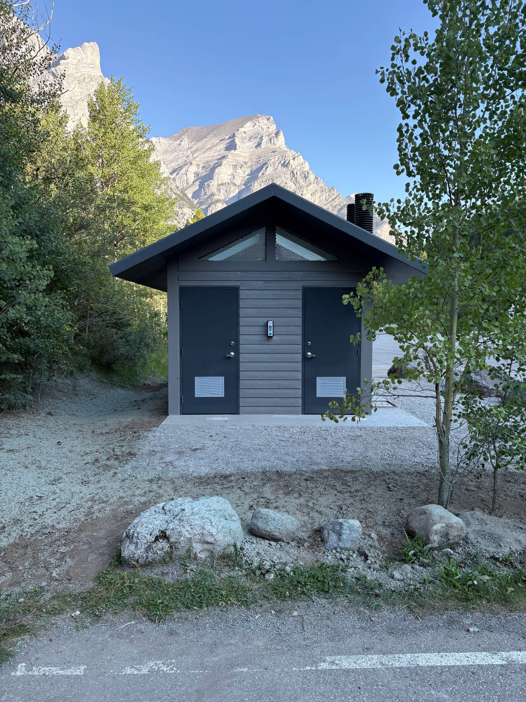 Front of a new vault toilet building with twin doors and rock landscaping below a mountain peak at Kananaskis Region Vault Toilets, built by Premiere Builders