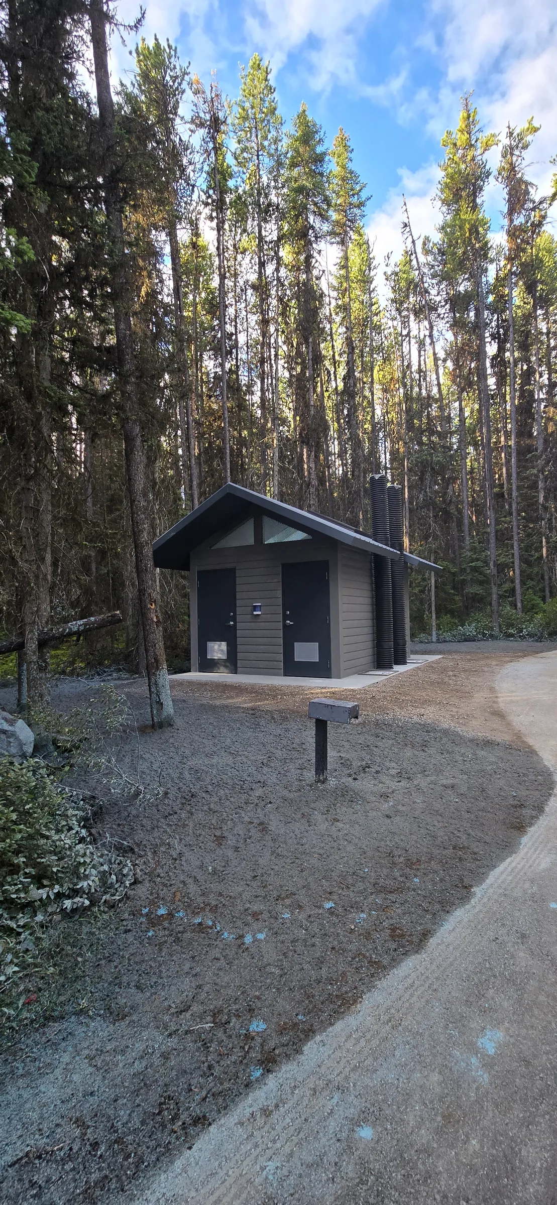 New vault toilet at the end of a gravel path surrounded by lodgepole pine forest at Kananaskis Region Vault Toilets, built by Premiere Builders