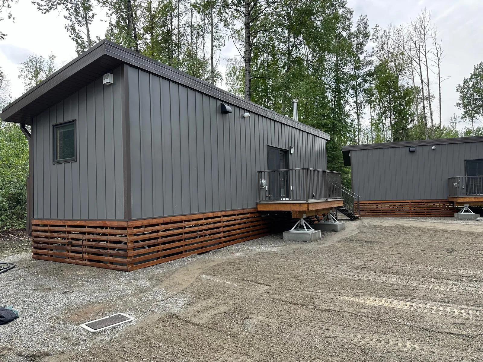Modular staff cabin with dark vertical metal siding, cedar skirting and a railed deck among boreal forest at Liard River Park Ranger Cabins, built by Premiere Builders