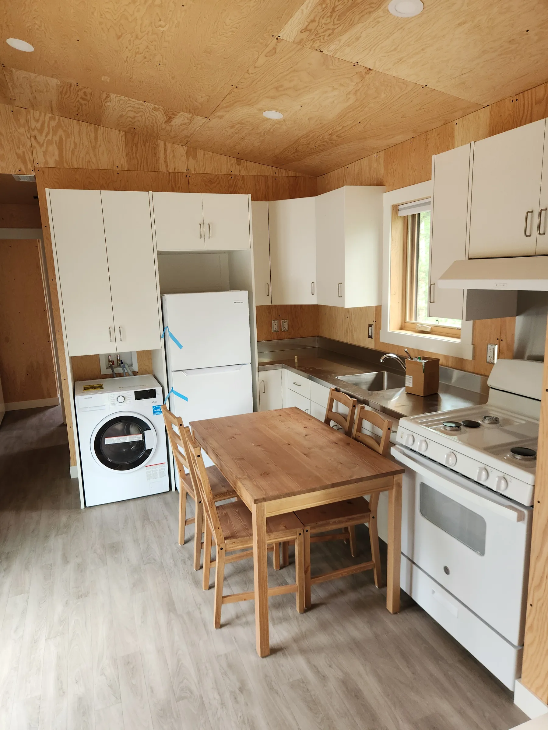 Cabin kitchen with white cabinets, wood-panelled walls, stacked laundry and a wood dining table at Liard River Park Ranger Cabins, built by Premiere Builders