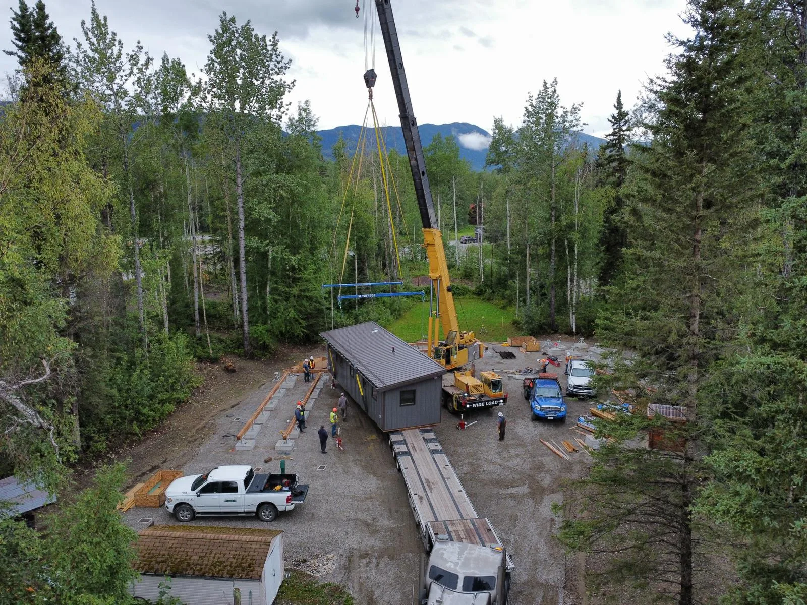 Aerial view of a crane setting a modular cabin into place at a forested camp site with mountains behind at Liard River Park Ranger Cabins, built by Premiere Builders