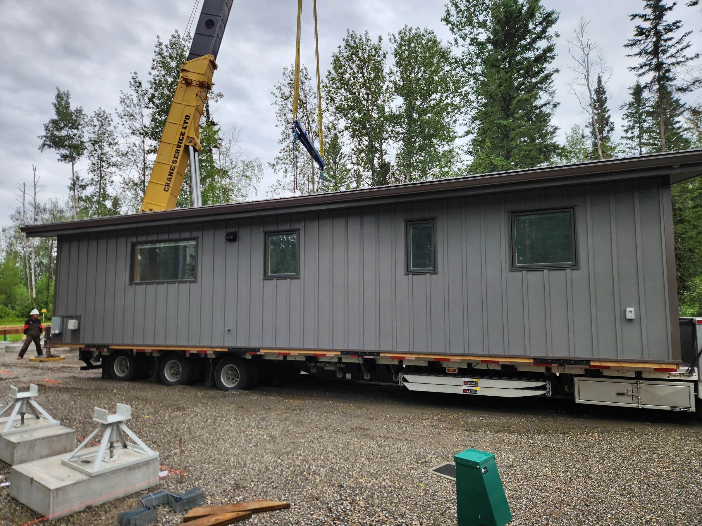 Crane positioning a finished modular cabin delivered on a flatbed trailer at a forested site at Liard River Park Ranger Cabins, built by Premiere Builders