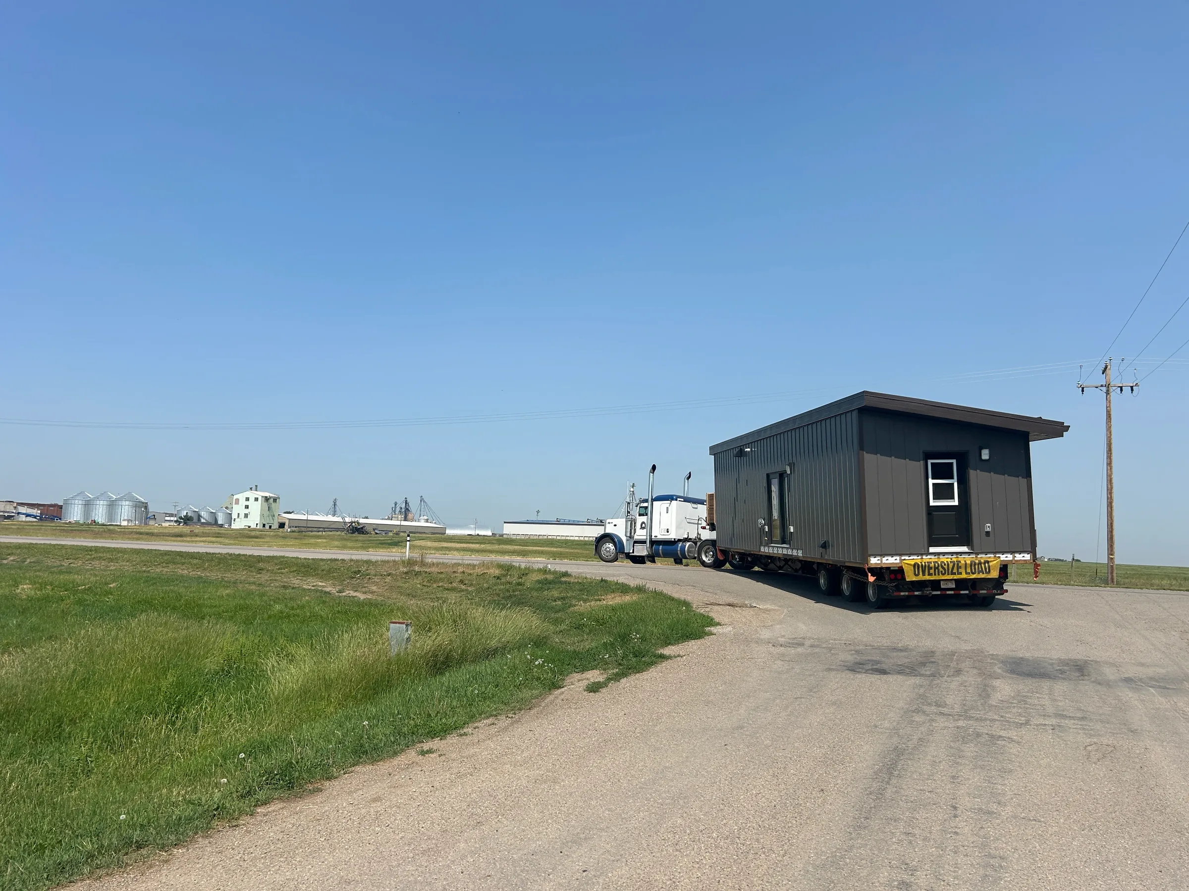 Completed modular cabin loaded on an oversize-load trailer for transport along a prairie road at Liard River Park Ranger Cabins, built by Premiere Builders