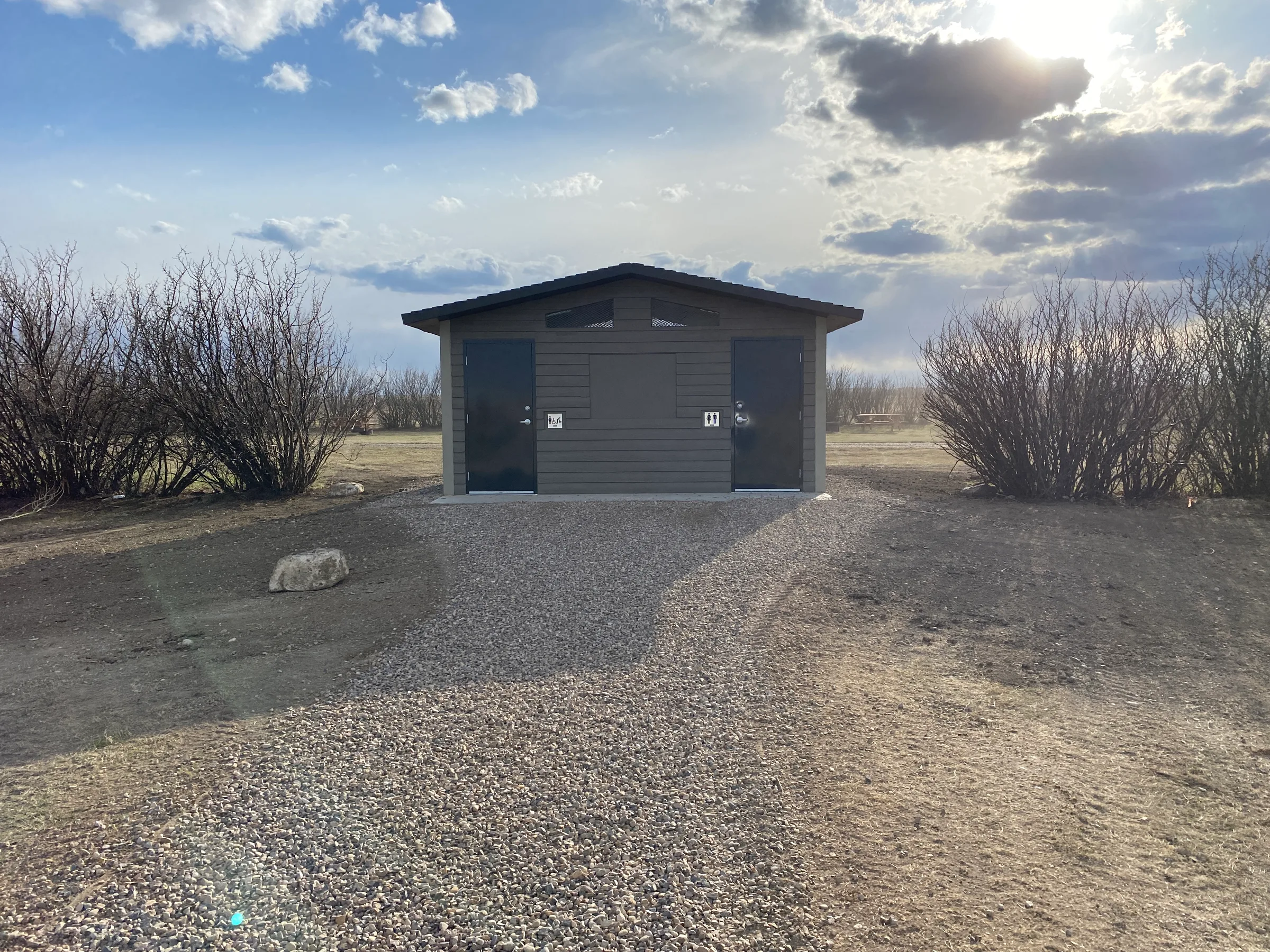 Front of a completed vault toilet at the end of a gravel walkway framed by shrubs at Lundbreck Falls and Lake McGregor Vault Toilets, built by Premiere Builders