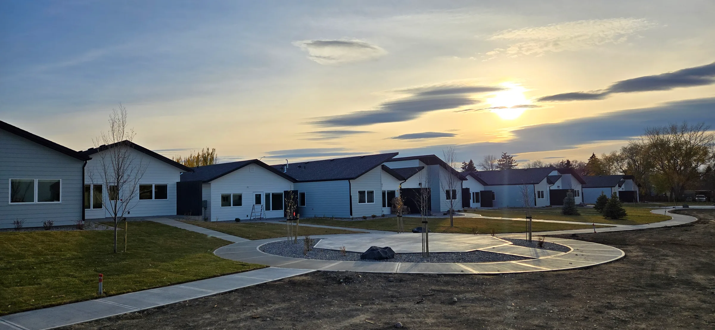 Seniors cottages around a landscaped common area with a curved walkway and round patio at sunset at Porcupine Hills Lodge, built by Premiere Builders