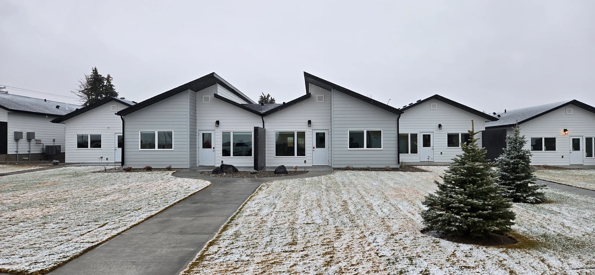 Completed fourplex with a walkway and young spruce trees on a light dusting of snow at Porcupine Hills Lodge, built by Premiere Builders